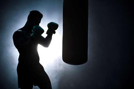 Silhouette of an unrecognizable boxer man sparring with a punching bag in a dark gym. Male athlete working out in preparation for his upcoming fight. Young martial artist training and practicing